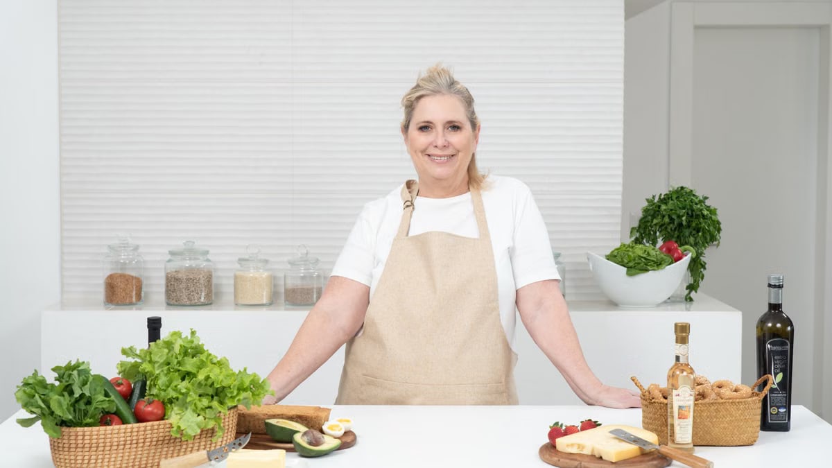 A mature woman in an apron stands at a kitchen counter with fresh produce and cheese, ready for cooking.